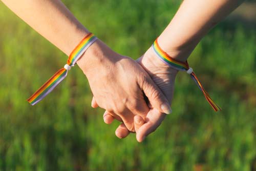 Zwei Hände, die ineinander verschränkt sind. Am Handgelenk tragen sie regenbogenfarbene Armbänder.